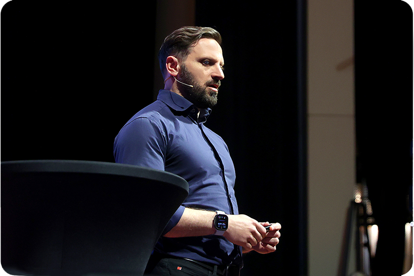 Fatmir talar på ett företagsevent i blå skjorta på mörk scen